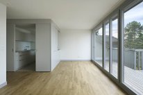 Ausschnitt aus leerer Wohnung. Auf rechter Seite ist eine Fensterfront zu sehen und in der linken Ecke kleiner Einblick in die Küche.