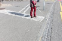 Ein breites Trottoire, auf dem an einem Strassenübergang eine Person mit Blindenstock steht.