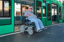 Ein Mann in einem grossen elektrischen Rollstuhl, der aus einem Tram fährt.